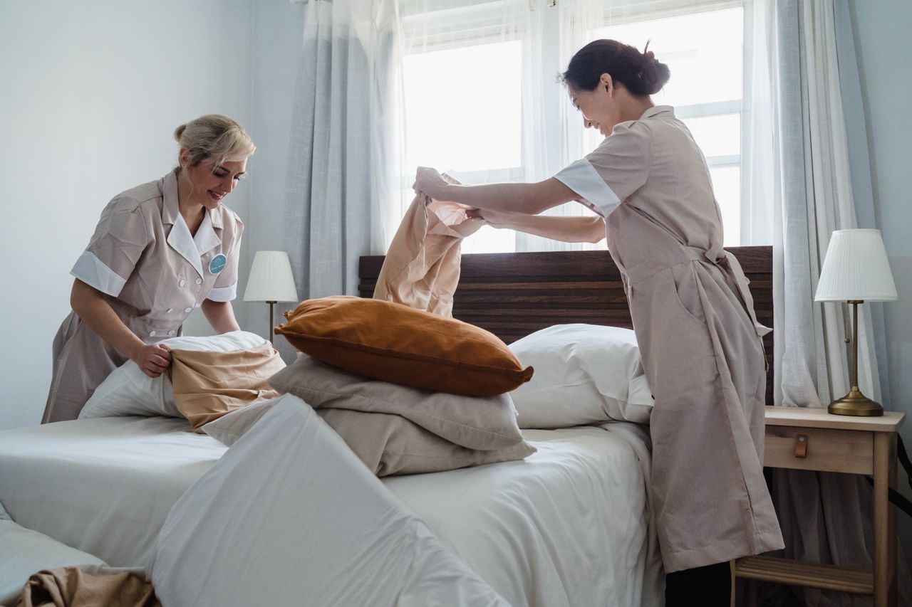 services-01 Deux employées d’entretien en uniforme refaisant le lit d’une chambre d’hôtel avec du linge propre et des oreillers frais.